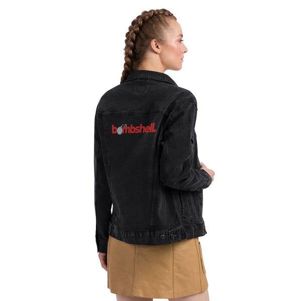 A young woman confidently poses in a  black denim 'Bombshell' jacket, styled with a grey t-shirt and a tan skirt. 